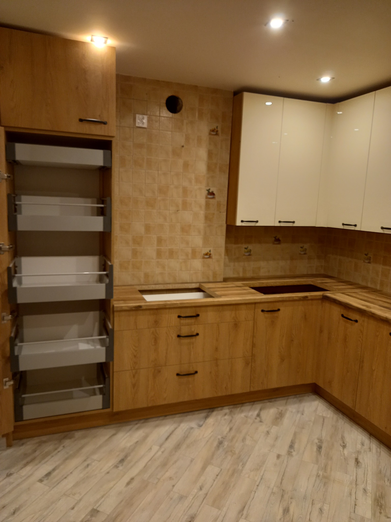 Jasna kuchnia z drewnianymi frontami i białymi szafkami wiszącymi, z otwartą szafką cargo z wysuwanymi półkami, płytki ceramiczne na ścianie i drewnopodobna podłoga.