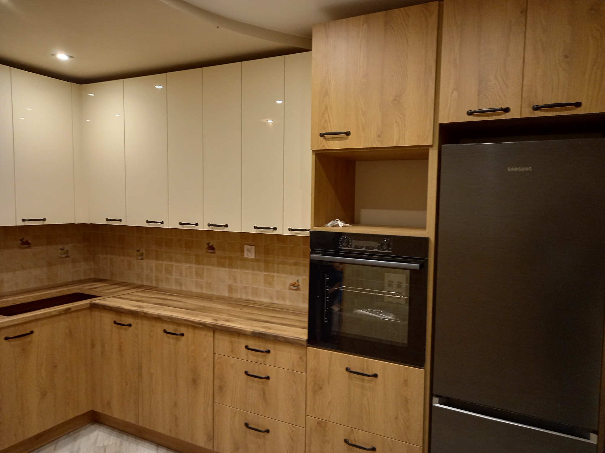 Jasna kuchnia z drewnianymi frontami szafek dolnych i białymi górnymi, z wbudowanym piekarnikiem i lodówką Samsung, widoczna płytka ceramiczna na ścianie.