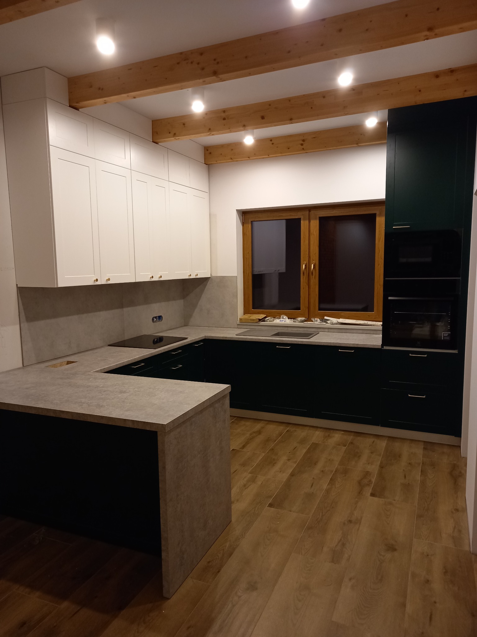 Nowoczesna kuchnia z białymi górnymi szafkami, ciemnozielonymi dolnymi szafkami i blatem w odcieniach szarości, z drewnianymi belkami stropowymi i oknem w drewnianej ramie.