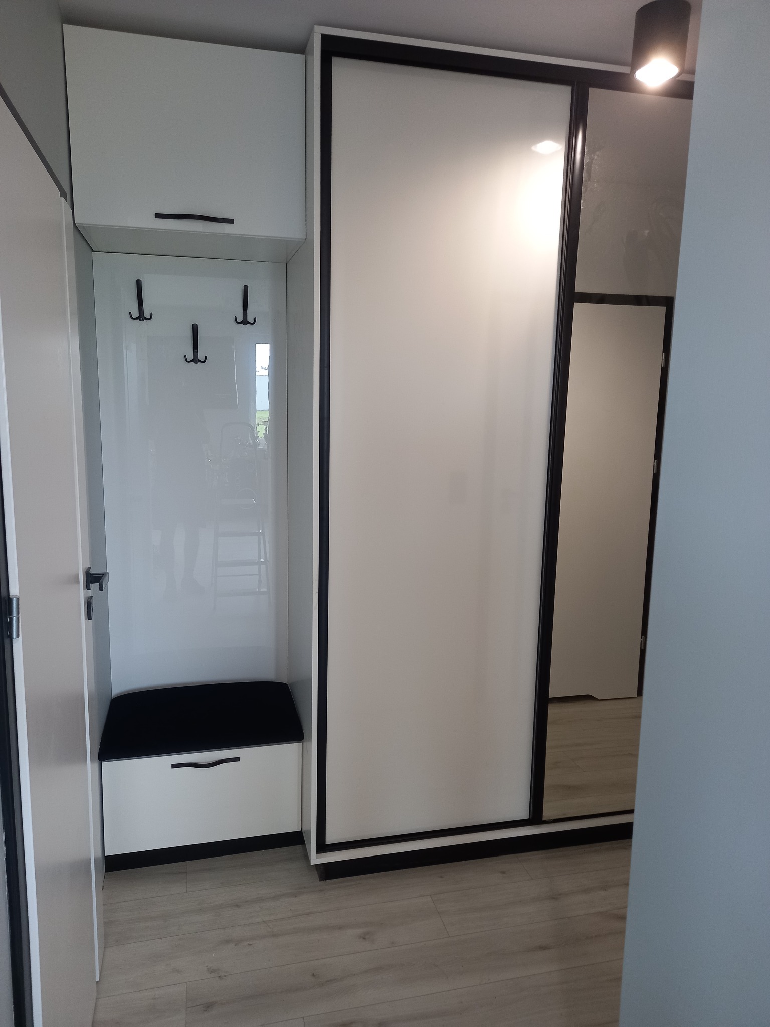 Biała szafa przesuwna z czarnymi obramowaniami i wbudowanym siedziskiem z wieszakami w przedpokoju, widoczny fragment podłogi z paneli.