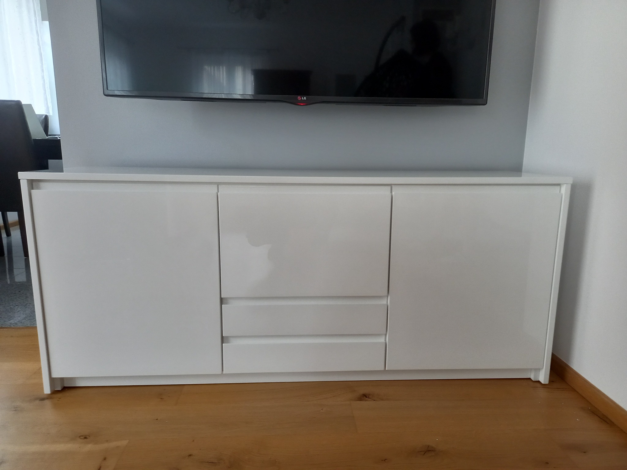 Biała, lakierowana komoda RTV z trzema szufladami i dwojgiem drzwi, stojąca na drewnianej podłodze, pod wiszącym telewizorem LG.