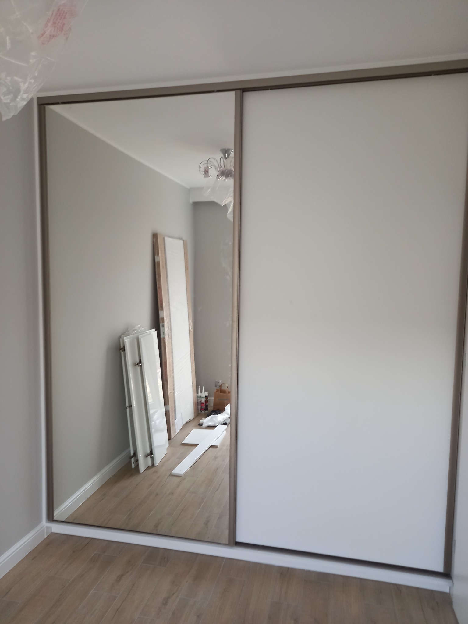 Wnętrze pokoju z zamontowaną szafą przesuwną z lustrzanym frontem, widoczne elementy montażowe i akcesoria, sugerujące trwający proces instalacji.