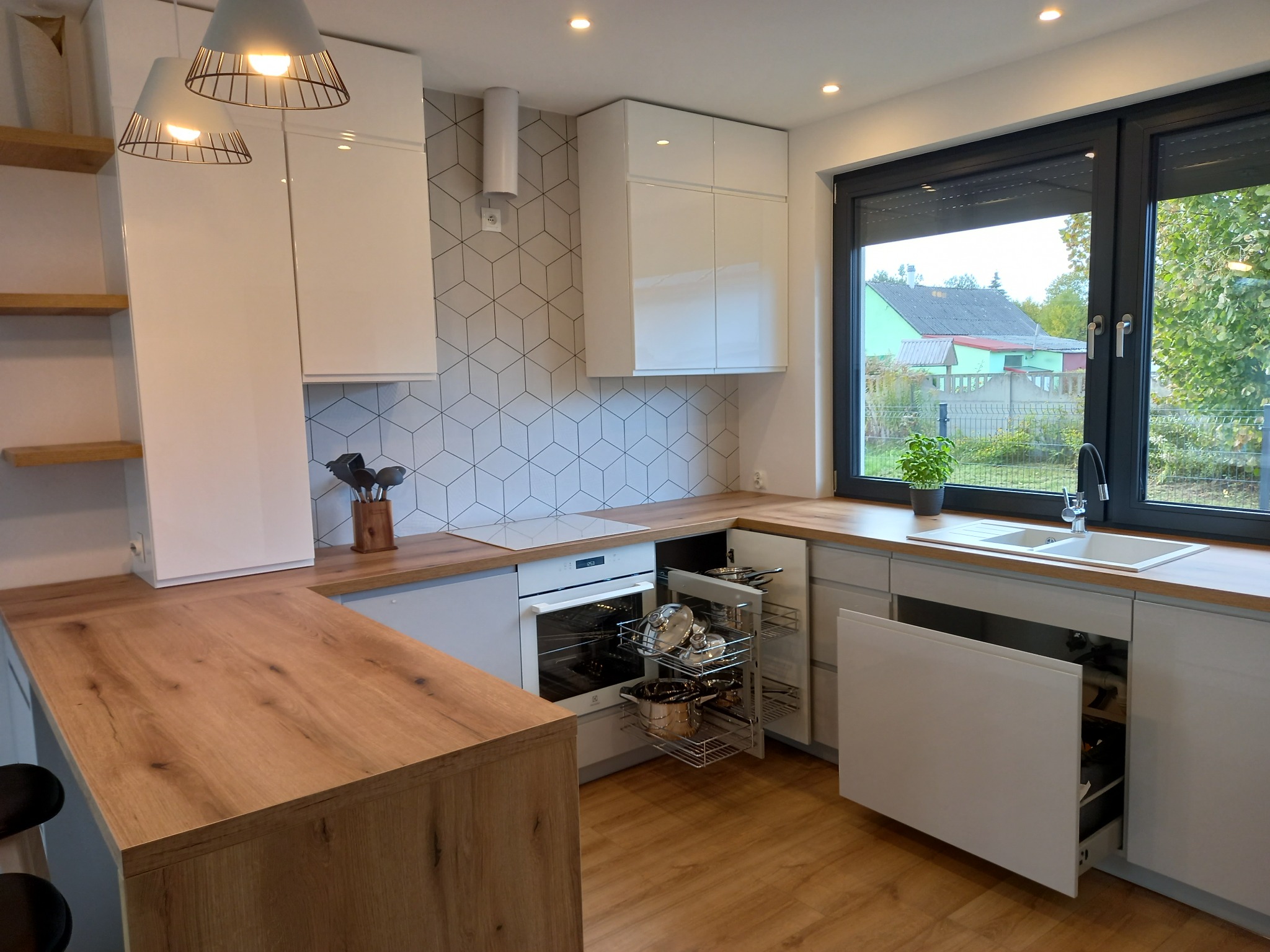 Nowoczesna kuchnia z białymi, lakierowanymi frontami, drewnianym blatem i wysuwanymi koszami na garnki, widok na okno z roletami zewnętrznymi i zieleń za nim.