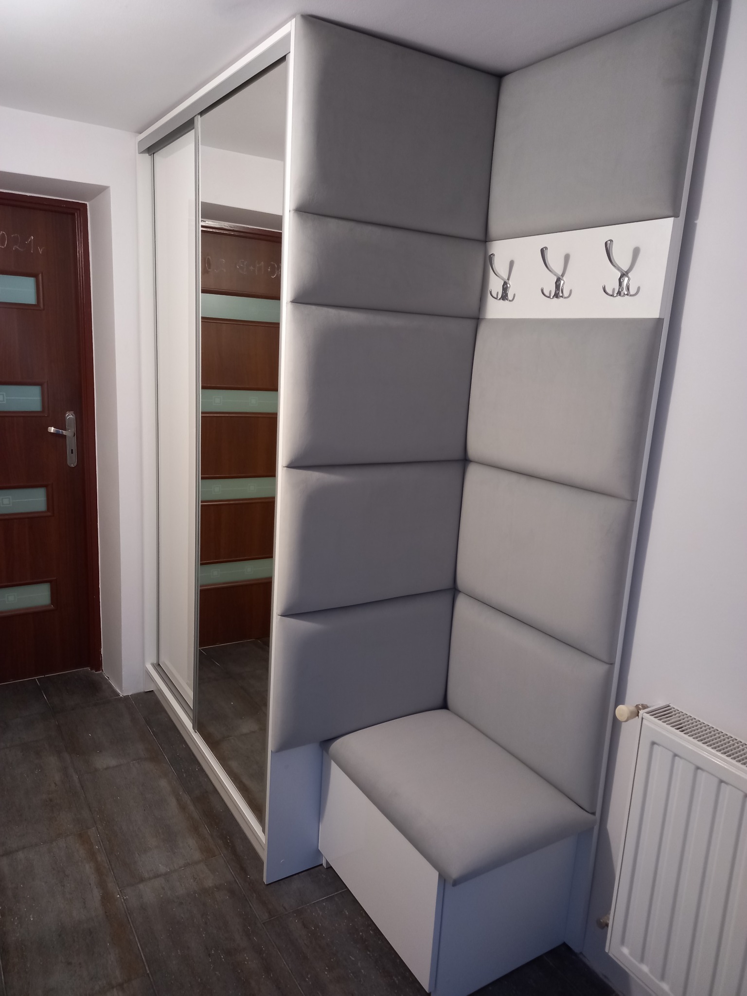Szara szafa wnękowa z lustrzanymi drzwiami przesuwnymi, obok tapicerowany panel ścienny z siedziskiem i wieszakami, tworzący kącik w przedpokoju.