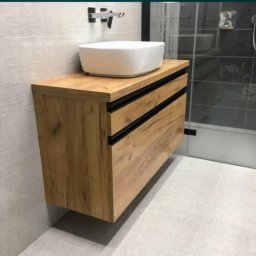 STOLARSTWO PIOTR GŁĄBOWSKI - Nowoczesna łazienka z drewnianą szafką pod umywalkę nablatową i szarą kabiną prysznicową w tle.