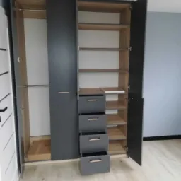 Szafa wnękowa z szarymi frontami, drewnianym wnętrzem i czterema szufladami, otwarte drzwi ukazują półki, widoczna klimatyzacja sufitowa.