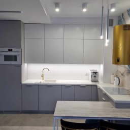 STOLARSTWO PIOTR GŁĄBOWSKI - Nowoczesna kuchnia w odcieniach szarości z złotymi akcentami, minimalistyczna zabudowa, designerski okap w kolorze złota na tle ściany z motywem akwareli, oświetlenie punktowe i wiszące lampy rurowe.