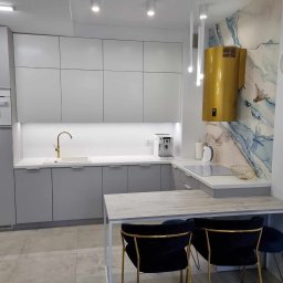 STOLARSTWO PIOTR GŁĄBOWSKI - Nowoczesna szara kuchnia z białym blatem, złotą armaturą i oryginalnym złotym okapem na tle ściany z motywem akwareli, uzupełniona czarnymi krzesłami z złotymi nogami.