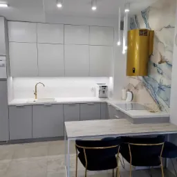 Nowoczesna szara kuchnia z białym blatem, złotą armaturą i oryginalnym złotym okapem na tle ściany z motywem akwareli, uzupełniona czarnymi krzesłami z złotymi nogami.