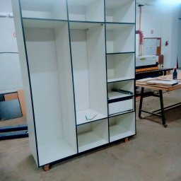 STOLARSTWO PIOTR GŁĄBOWSKI - Biała szafa z czarnymi obramowaniami w trakcie montażu, widoczne półki, szuflady i narzędzia na podłodze w warsztacie.