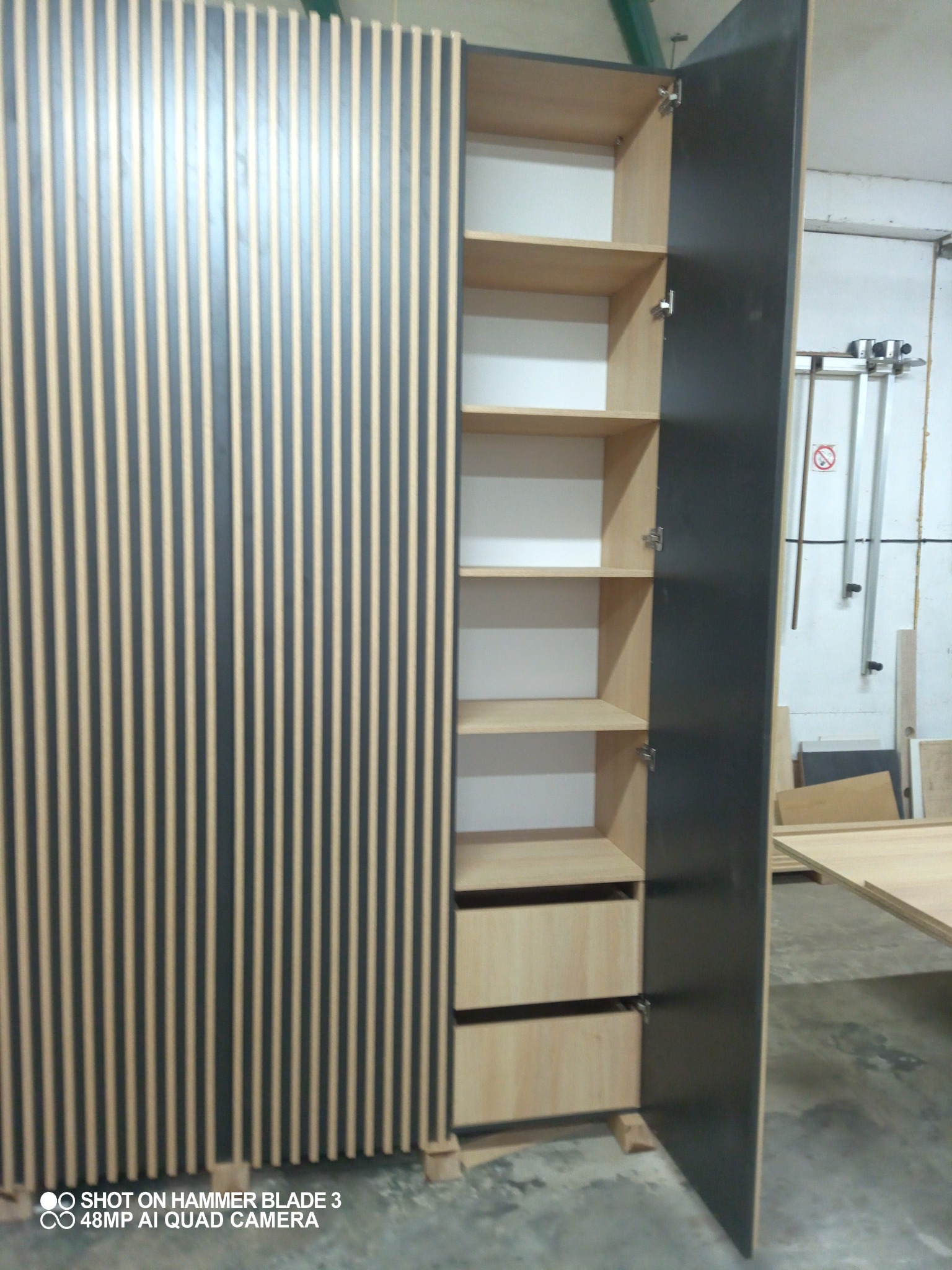 Otwarta szafa z drewnianymi półkami i szufladami, front z pionowymi panelami w kolorze grafitowym i jasnego drewna, widoczne zawiasy i fragment warsztatu w tle.