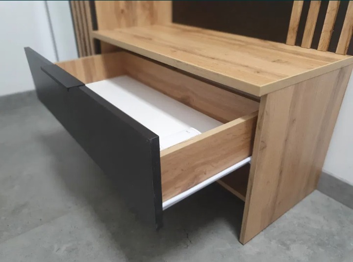 Otwarta szuflada z ciemnym frontem i jasnym wnętrzem w szafce z drewnopodobną okleiną, widoczny biały prowadnik szuflady.