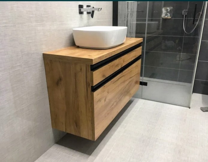 Nowoczesna łazienka z drewnianą szafką pod umywalkę nablatową i szarą kabiną prysznicową w tle.