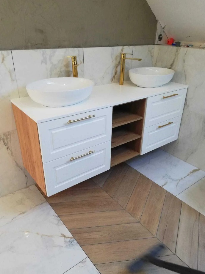 Nowoczesna szafka łazienkowa z dwoma białymi umywalkami nablatowymi i złotą armaturą, białe fronty szuflad z frezowaniem i złotymi uchwytami, drewniane półki, marmurowe płytki ścienne i podłoga...