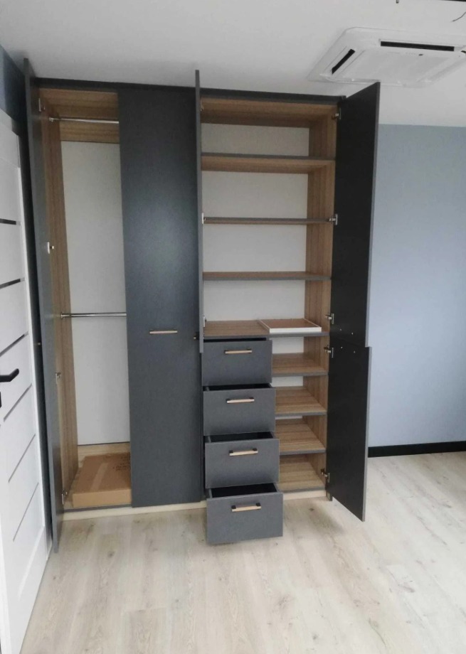 Szafa wnękowa z szarymi frontami, drewnianym wnętrzem i czterema szufladami, otwarte drzwi ukazują półki, widoczna klimatyzacja sufitowa.