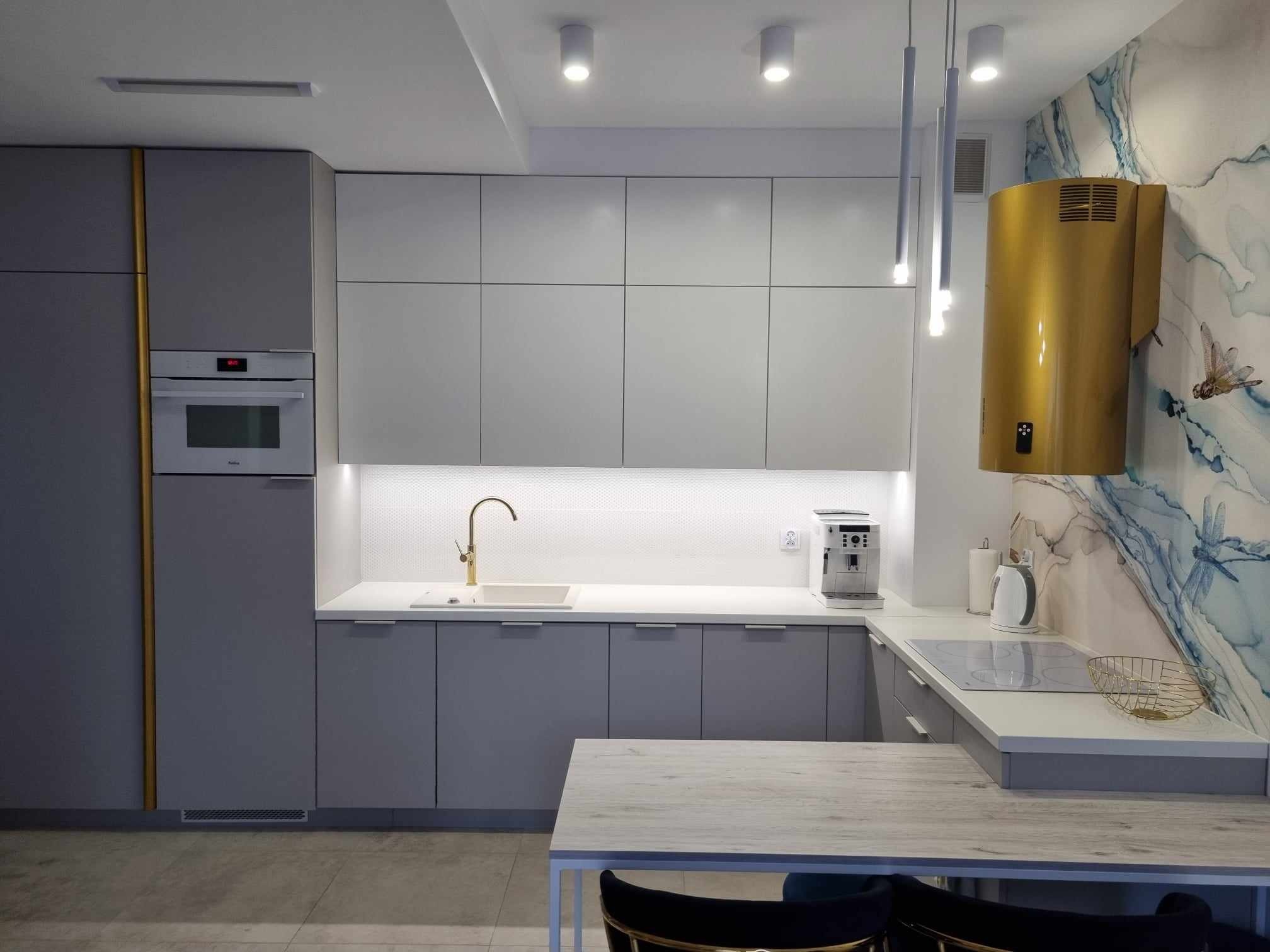 Nowoczesna kuchnia w odcieniach szarości z złotymi akcentami, minimalistyczna zabudowa, designerski okap w kolorze złota na tle ściany z motywem akwareli, oświetlenie punktowe i wiszące lampy rurowe.