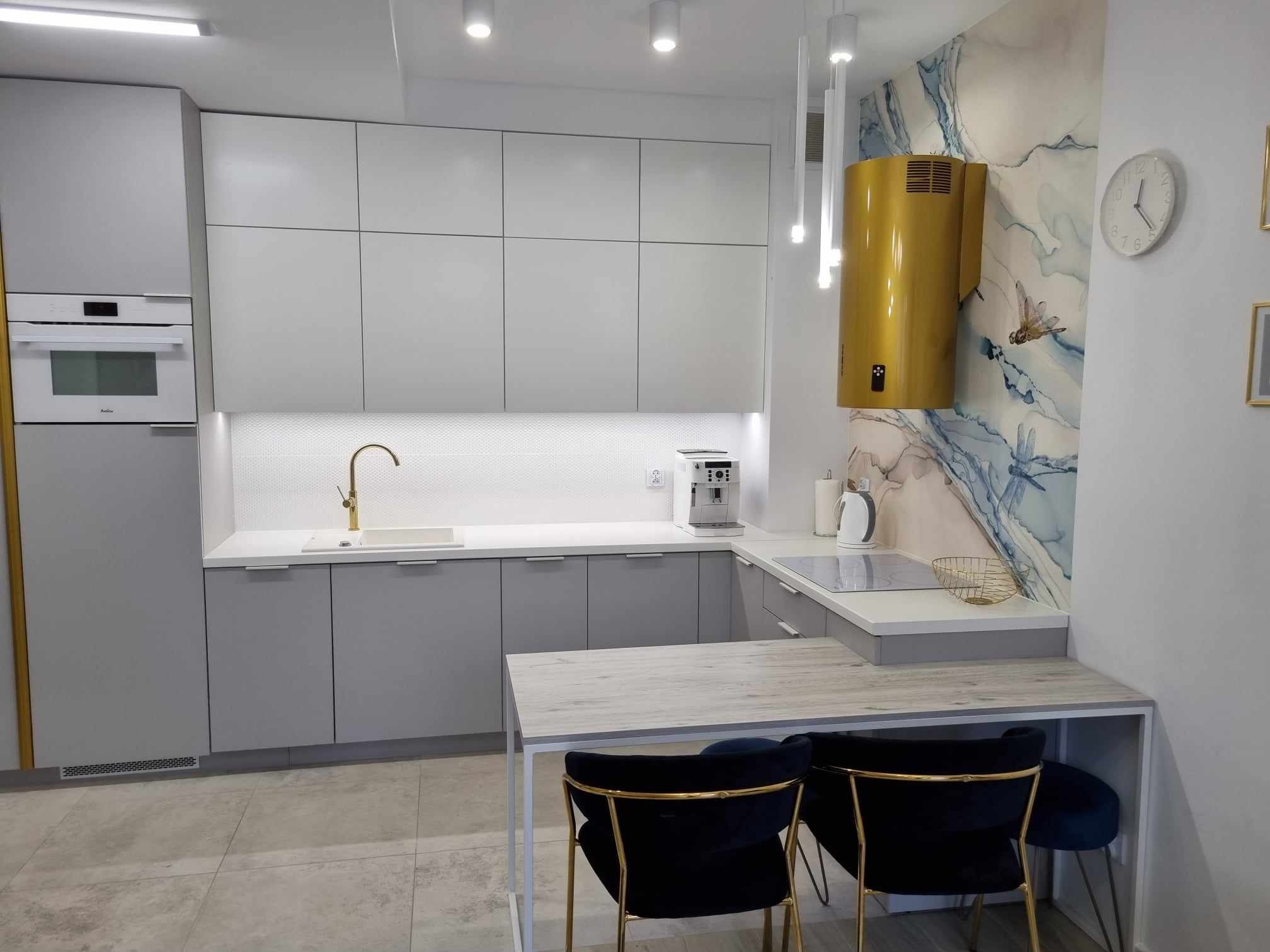 Nowoczesna szara kuchnia z białym blatem, złotą armaturą i oryginalnym złotym okapem na tle ściany z motywem akwareli, uzupełniona czarnymi krzesłami z złotymi nogami.