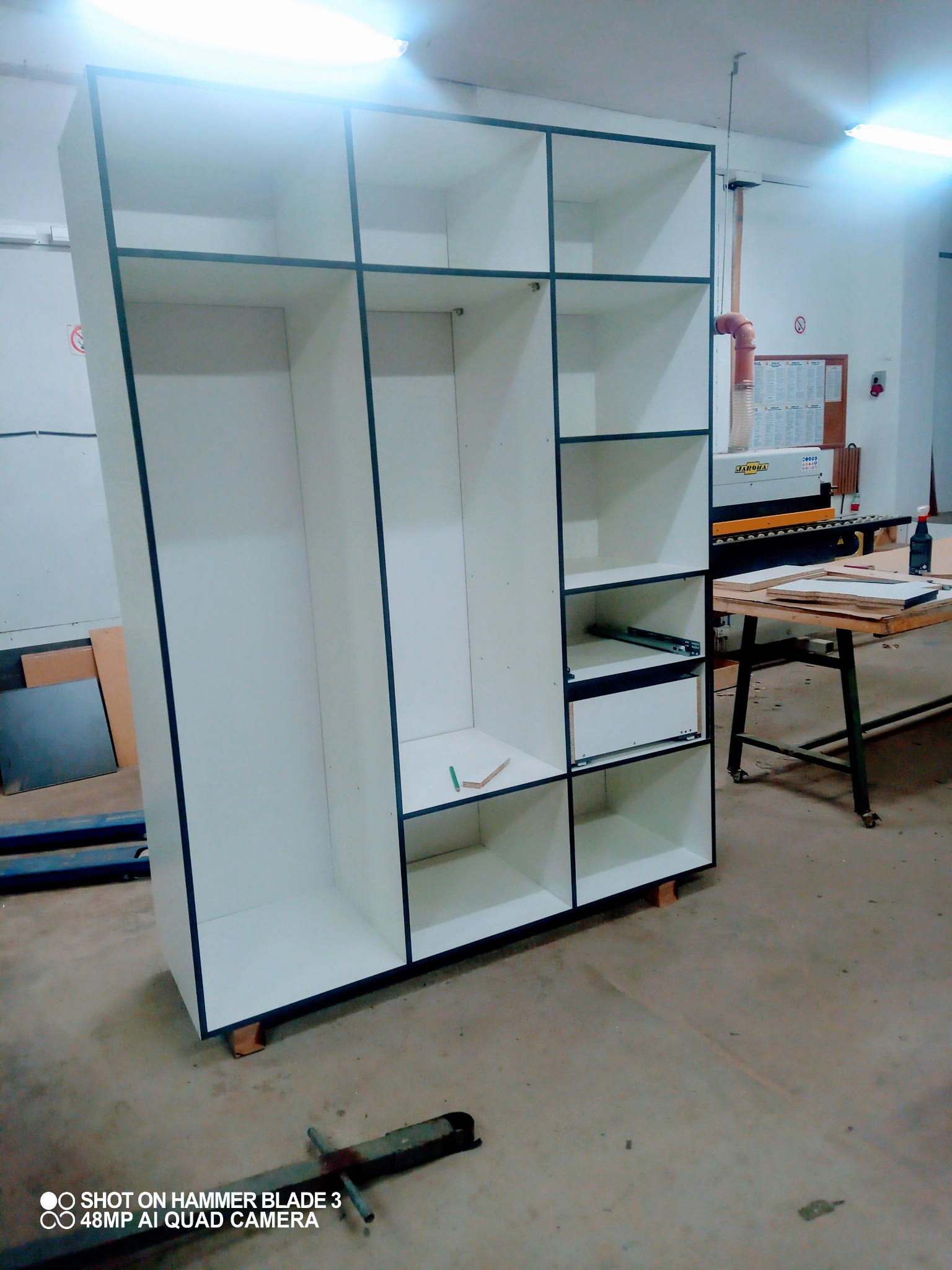 Biała szafa z czarnymi obramowaniami w trakcie montażu, widoczne półki, szuflady i narzędzia na podłodze w warsztacie.