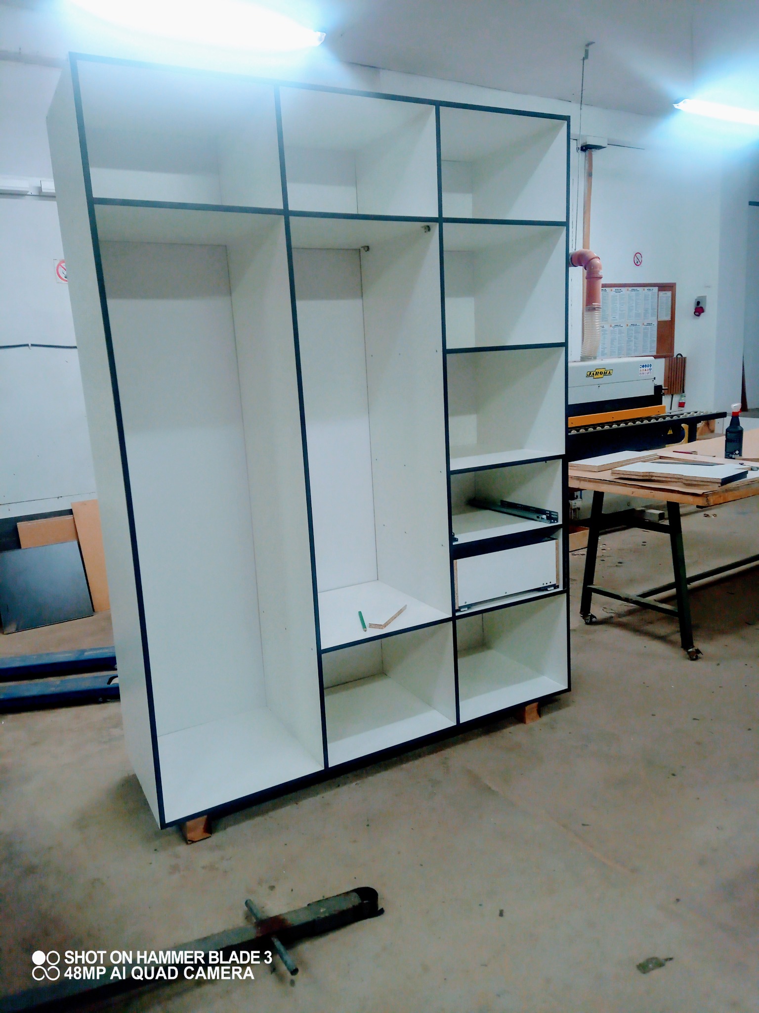 Wnętrze warsztatu stolarskiego z widokiem na białą, częściowo zmontowaną szafę z czarnymi obramowaniami, postawioną na drewnianych klockach, z widoczną piłą stołową w tle.