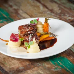 Elegancko podany stek z marchwią baby, puree ziemniaczanym i sosem na białym talerzu, na tle rustykalnego drewnianego stołu.