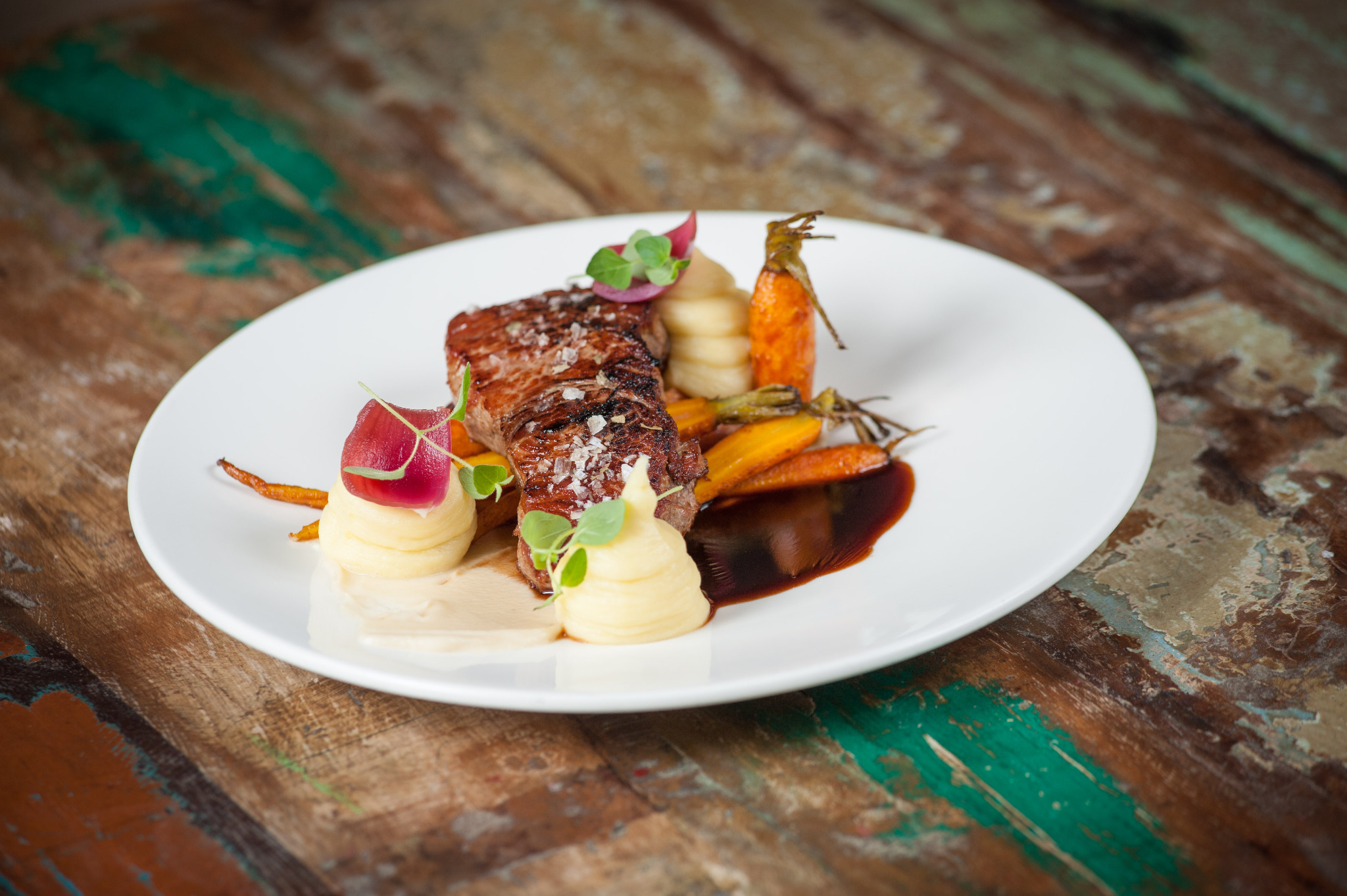 Elegancko podany stek z marchwią baby, puree ziemniaczanym i sosem na białym talerzu, na tle rustykalnego drewnianego stołu.