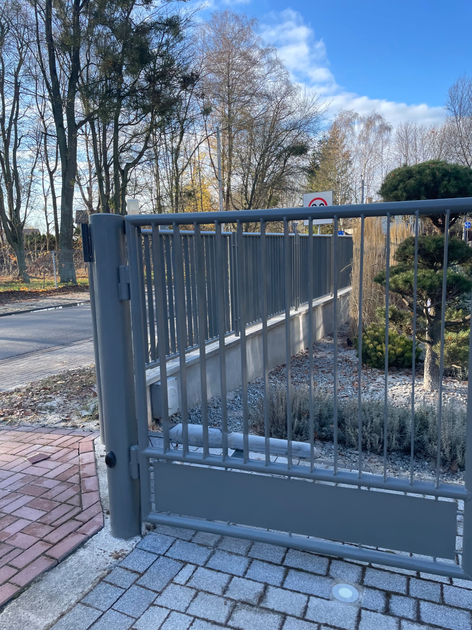 Nowoczesna, szara brama posesyjna z pionowymi prętami, widoczna z perspektywy chodnika wyłożonego kostką, z roślinnością i drzewami w tle oraz znakiem drogowym.