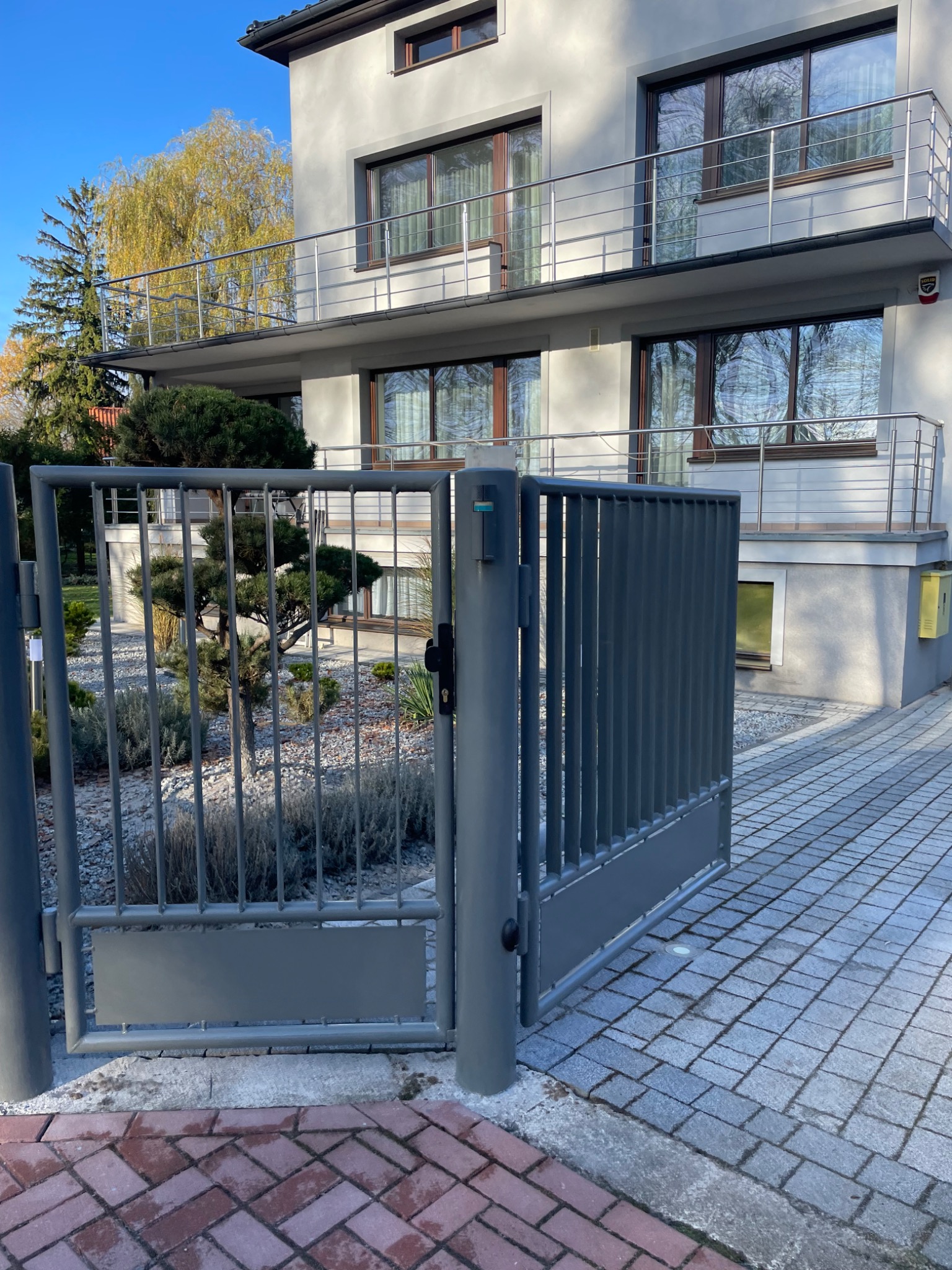 Szara, metalowa brama posesji w Elblągu, z widokiem na dwupiętrowy dom z balkonami i starannie utrzymany ogród.