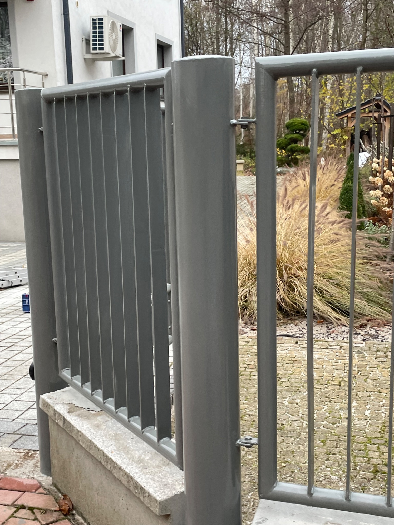 Szara, metalowa brama z pionowymi prętami, widok z bliska na słupki i fragment ogrodzenia, w tle trawa ozdobna, drzewa i element architektury ogrodowej.