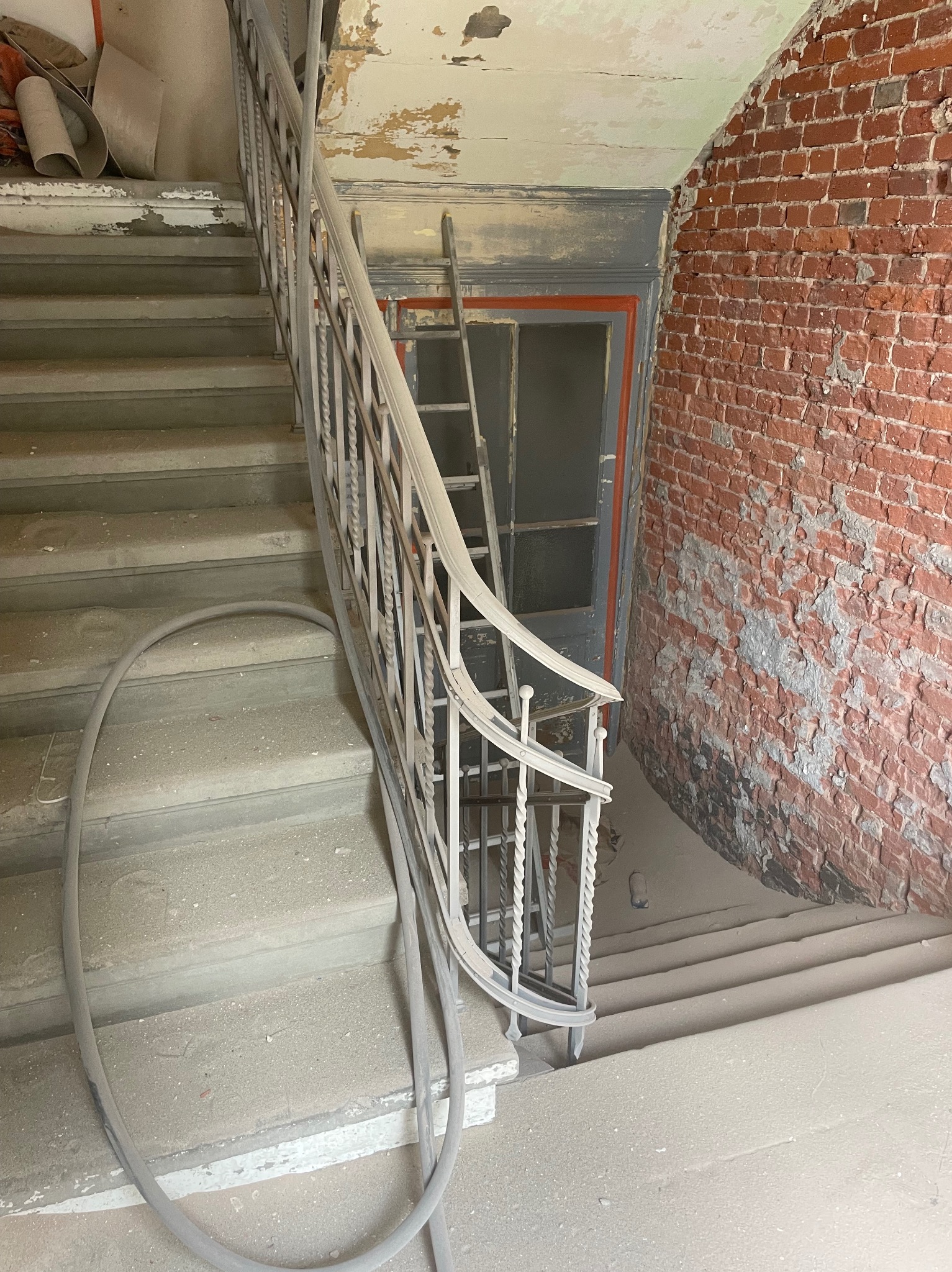 Klatka schodowa w trakcie renowacji: schody i balustrada pokryte warstwą pyłu, odsłonięta ceglana ściana z resztkami farby, drabina oparta o futrynę drzwi.