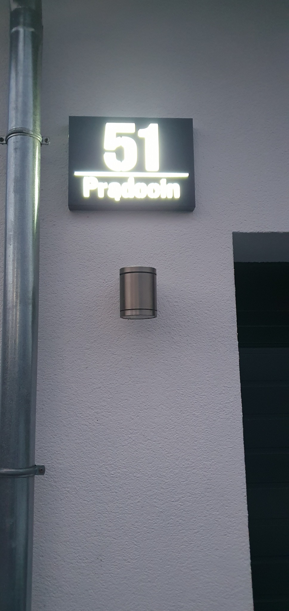Podświetlany numer domu '51' z nazwą ulicy 'Prądooin' zamontowany na elewacji obok metalowej rury spustowej i lampy zewnętrznej w cylindrycznej obudowie.