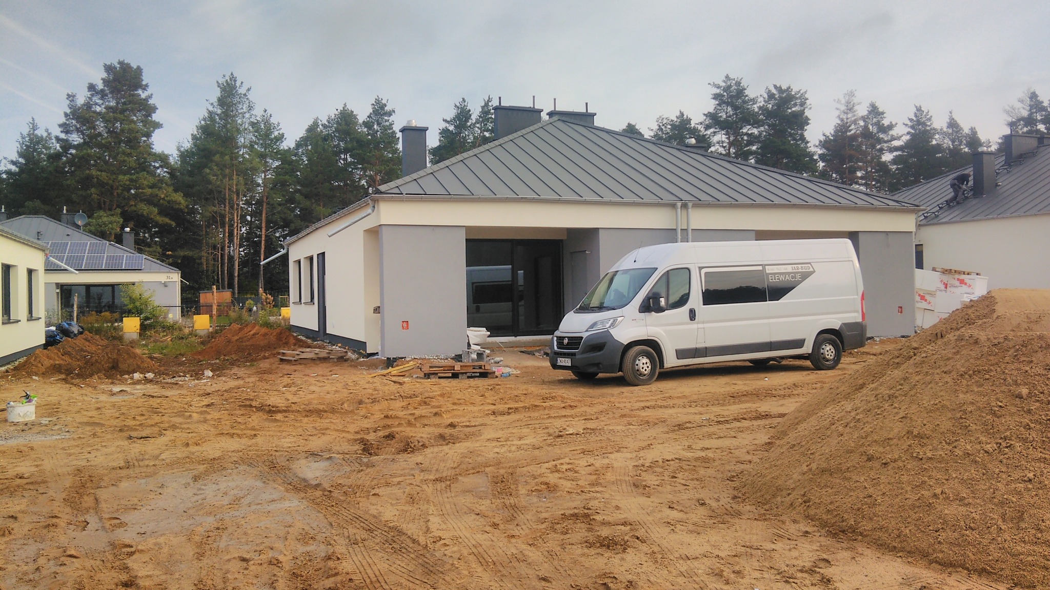 Nowoczesny dom jednorodzinny z szarą elewacją i ciemną stolarką okienną, widoczny biały samochód dostawczy z napisem 'Elewacje' w trakcie prac wykończeniowych, na pierwszym planie teren budowy...