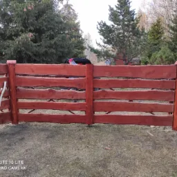 Czerwono-brązowe drewniane ogrodzenie z nieregularnymi krawędziami desek, widok na ogród z drzewami iglastymi i budynkiem w tle, częściowo zasłonięte przez sylwetkę osoby przy samochodzie.