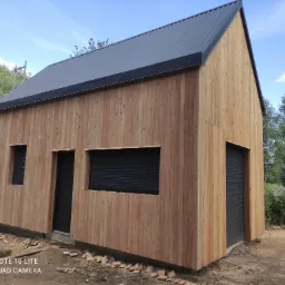 Surowy dom szkieletowy z desek z szarą blachą falistą na dachu, z czarnymi roletami okiennymi i drzwiami garażowymi, w trakcie wykończenia terenu wokół budynku.