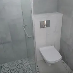 Nowoczesna łazienka w odcieniach szarości, z kabiną prysznicową ze szklanymi drzwiami, wiszącą toaletą i ozdobnymi płytkami podłogowymi w strefie prysznica.