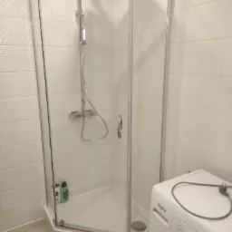 Narożna kabina prysznicowa z chromowanymi profilami i deszczownicą, widoczna w sąsiedztwie pralki w łazience z białymi płytkami o delikatnej strukturze.