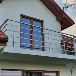 Balkon z prostą, metalową barierką na tle dwuspadowego dachu i okien w brązowych ramach.