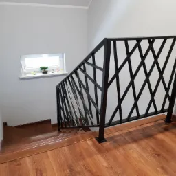 Schody wewnętrzne z drewnianymi stopniami i nowoczesną, czarną balustradą z motywem krzyżujących się ukośnych linii, widok z góry.