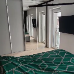 Sypialnia z łóżkiem w zielonej pościeli z geometrycznym wzorem, na ścianie duży obraz z motywem roślinnym i złota lampka, obok szafa przesuwna z lustrzanymi drzwiami, widok z okna na budowę.