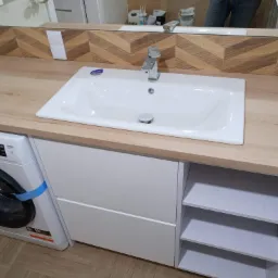 Łazienka z drewnianym blatem, białą umywalką i pralką obok szafki pod umywalką. W tle lustro i płytki ułożone w jodełkę.