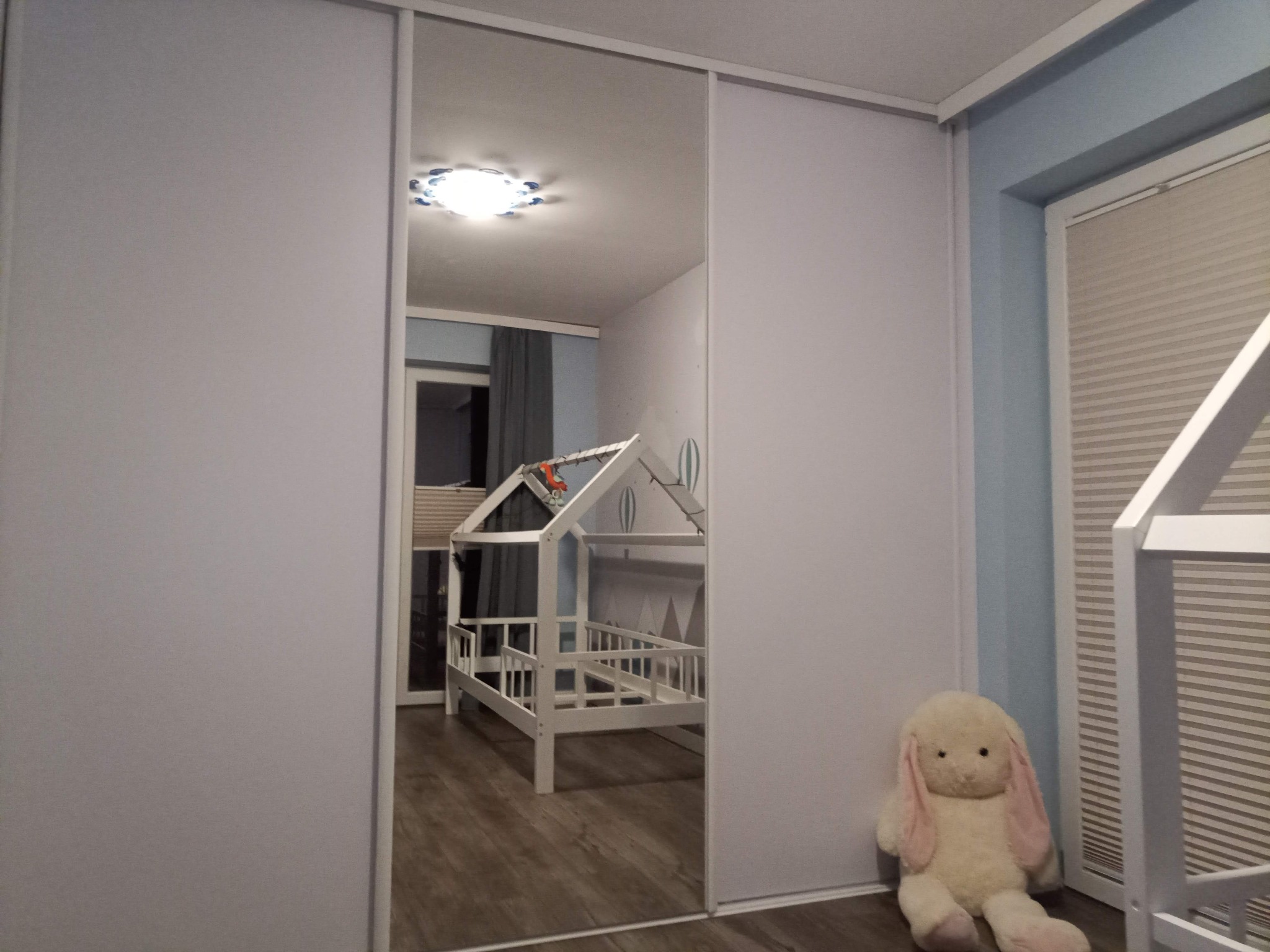 Biała szafa przesuwna z lustrzanymi drzwiami, odbijająca pokój dziecięcy z łóżkiem w kształcie domku i pluszowym królikiem siedzącym obok