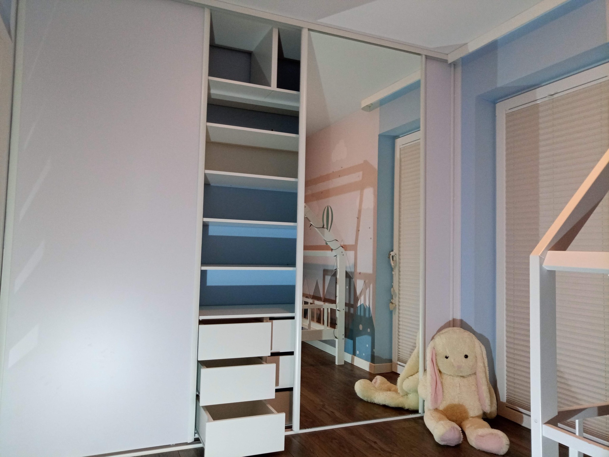 Biała szafa przesuwna z otwartymi szufladami w pokoju dziecięcym z łóżkiem piętrowym i pluszowym królikiem.