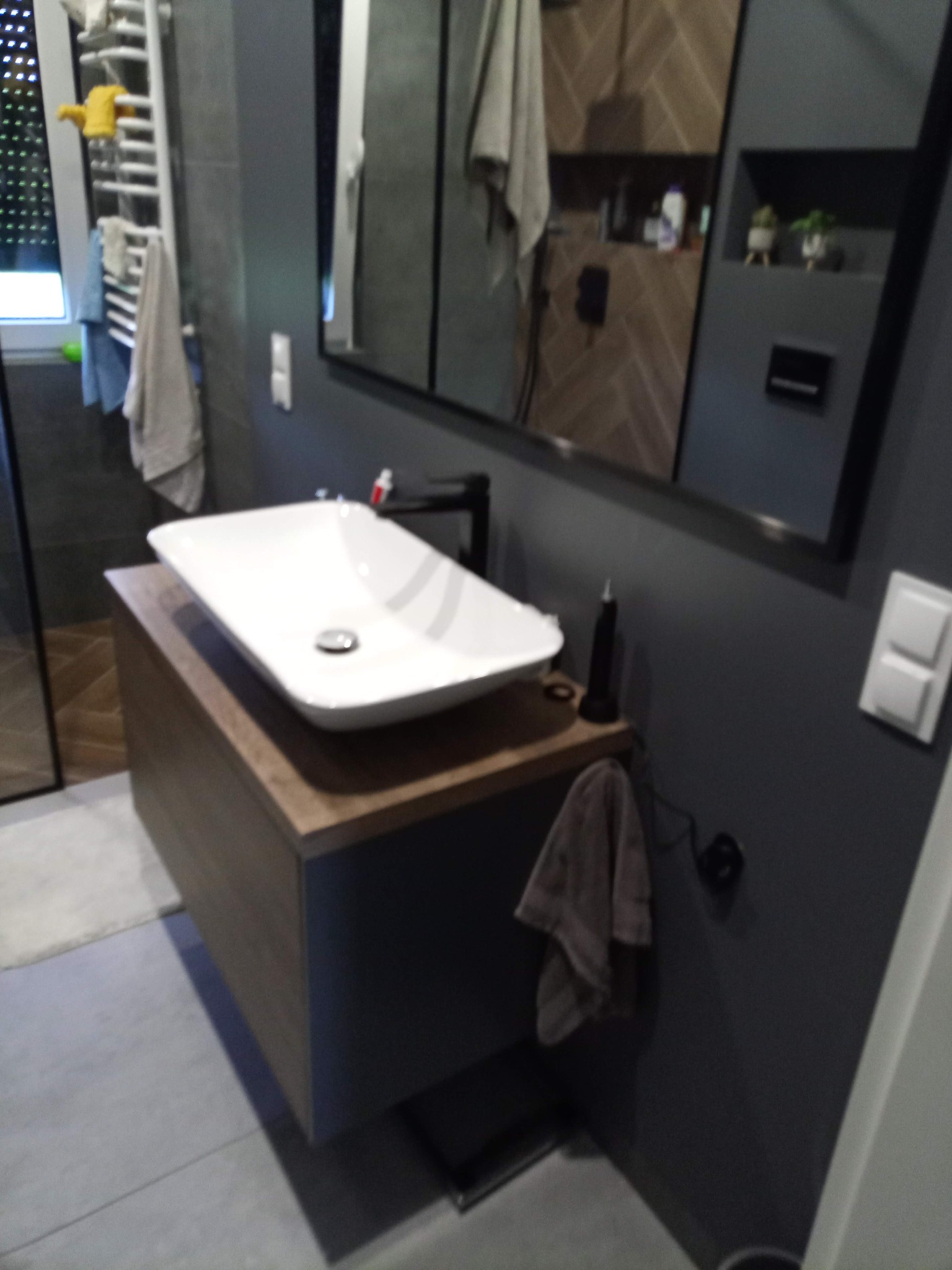 Nowoczesna łazienka z białą, prostokątną umywalką nablatową i czarną armaturą, zamontowaną na szafce z drewnianym blatem, widoczne szare ściany i lustro z odbiciem wnętrza łazienki.