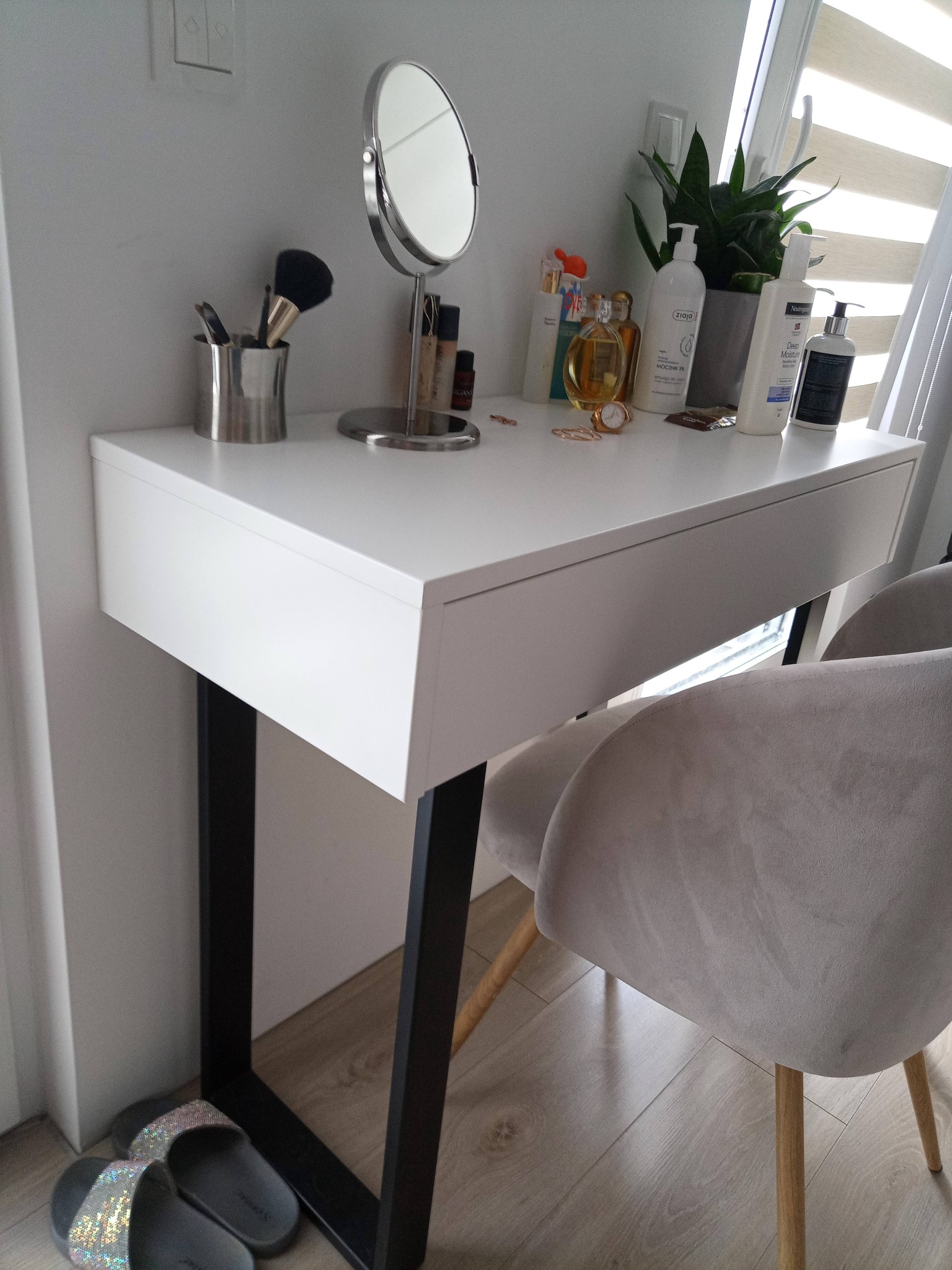 Biała toaletka z szufladą na czarnych nogach, okrągłe lustro kosmetyczne, pędzle do makijażu w srebrnym pojemniku, kosmetyki i roślina doniczkowa na blacie, szare welurowe krzesło i błyszczące...