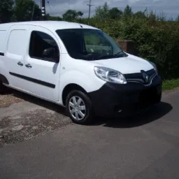 Biały samochód dostawczy Renault Kangoo zaparkowany na poboczu drogi, z widocznym logo Renault na masce i zielonymi krzewami w tle.