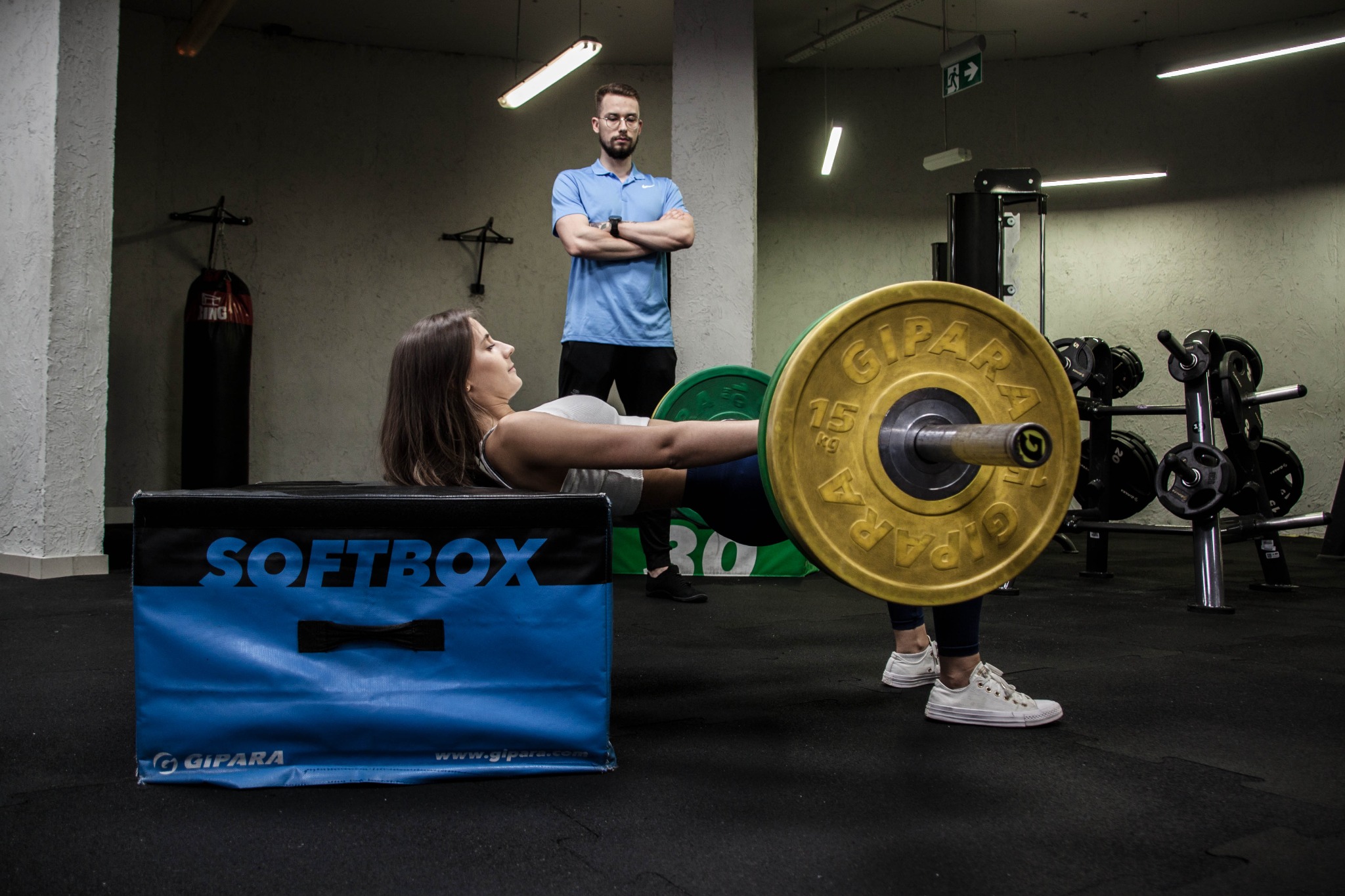 Kobieta wykonuje unoszenie bioder ze sztangą, opierając plecy na niebieskim boxie SOFTBOX w siłowni, pod nadzorem mężczyzny w okularach.