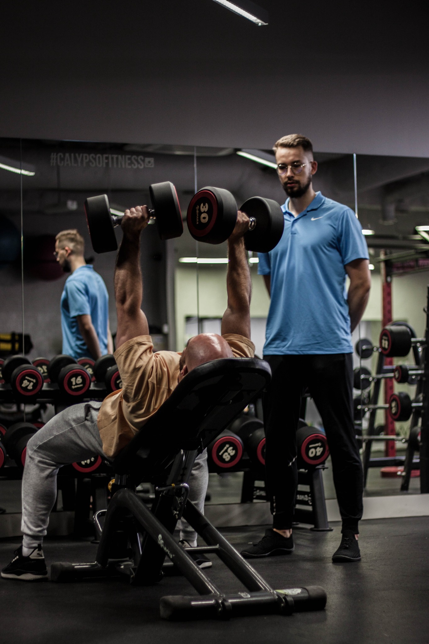 Mężczyzna na ławce skośnej wyciska hantle 30kg, nadzorowany przez mężczyznę w okularach w niebieskiej koszulce polo, siłownia Calypso Fitness w tle.