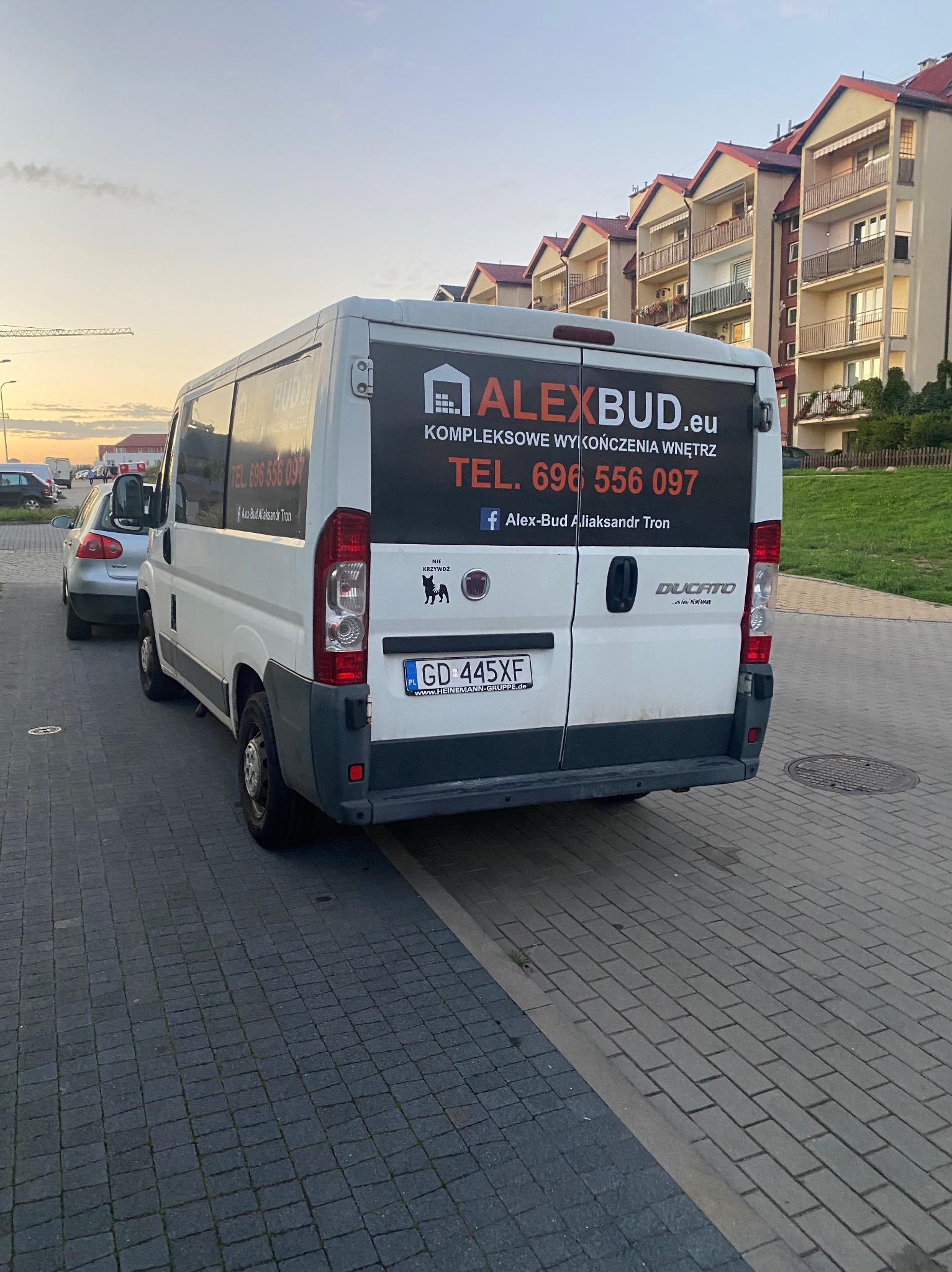 Biały bus marki Fiat Ducato z logo firmy 'ALEXBUD.eu Kompleksowe wykończenia wnętrz' zaparkowany na chodniku z kostki brukowej na tle bloku mieszkalnego.