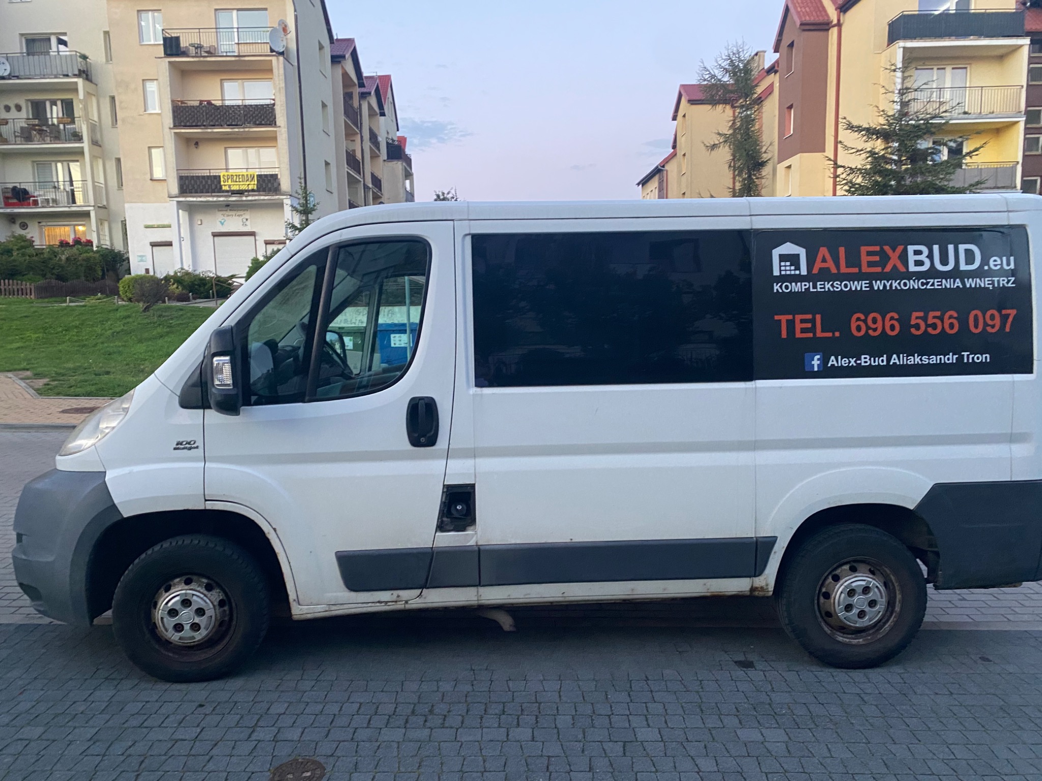 Biały bus dostawczy z reklamą firmy ALEXBUD.eu specjalizującej się w kompleksowym wykańczaniu wnętrz, zaparkowany na tle budynków mieszkalnych.