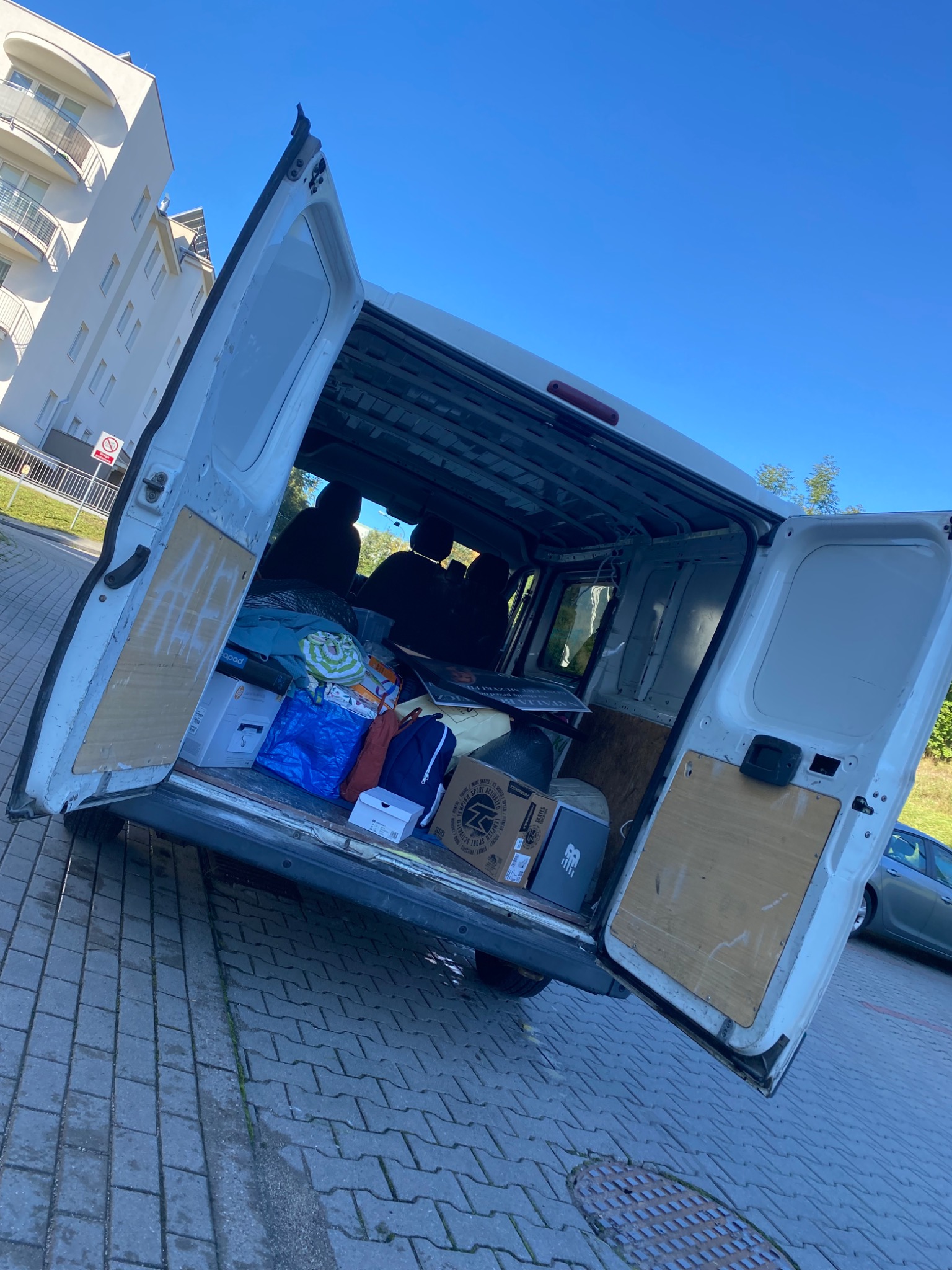 Otwarty biały furgon załadowany pudełkami, torbami i osobistymi rzeczami na tle budynku mieszkalnego i błękitnego nieba.