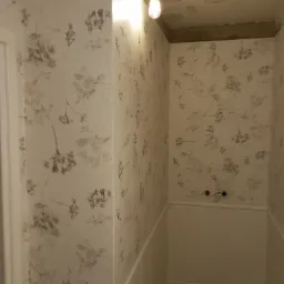 Wnętrze małej łazienki po odświeżeniu, z tapetą w motywy roślinne i niską boazerią, surowy sufit z żarówką bez klosza.